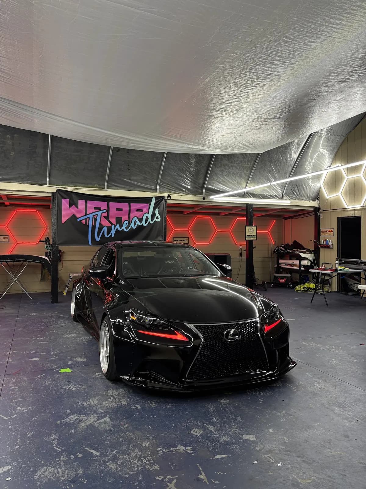 A black Lexus IS under the neon Wrap Threads banner in the studio, red hexagon lights on the wall behind it