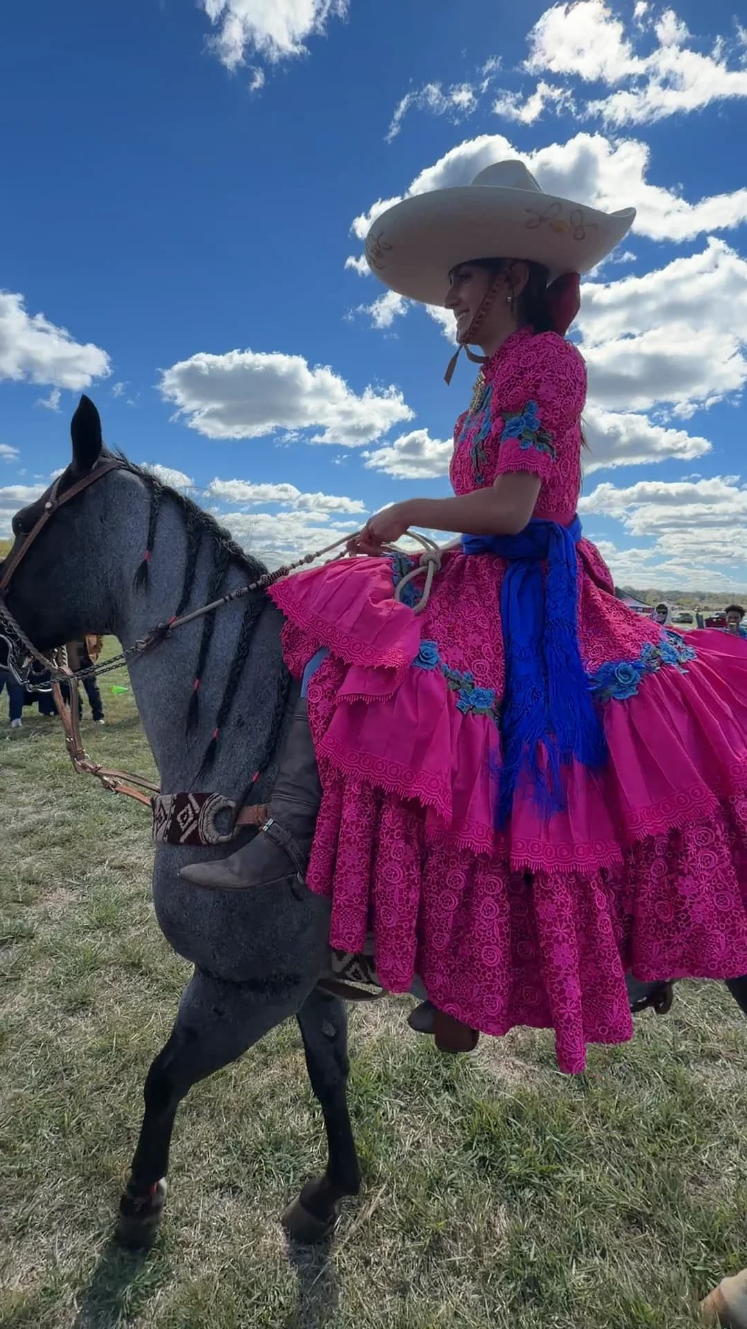 Jackie Jaramillo smiling in a wide charro hat and a pink escaramuza dress, riding sidesaddle on a blue roan horse