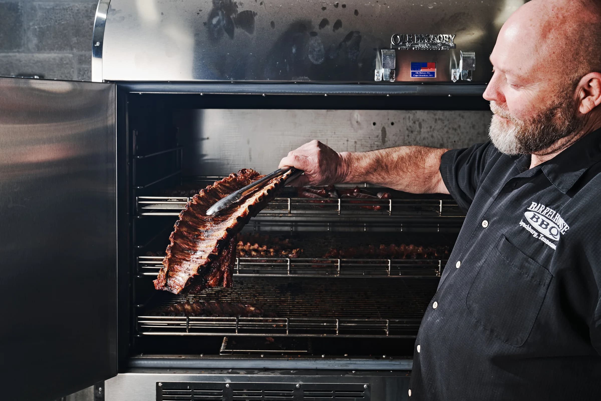 A pitmaster in a black Barrel House BBQ shirt lifts a rack of ribs from a stainless steel smoker