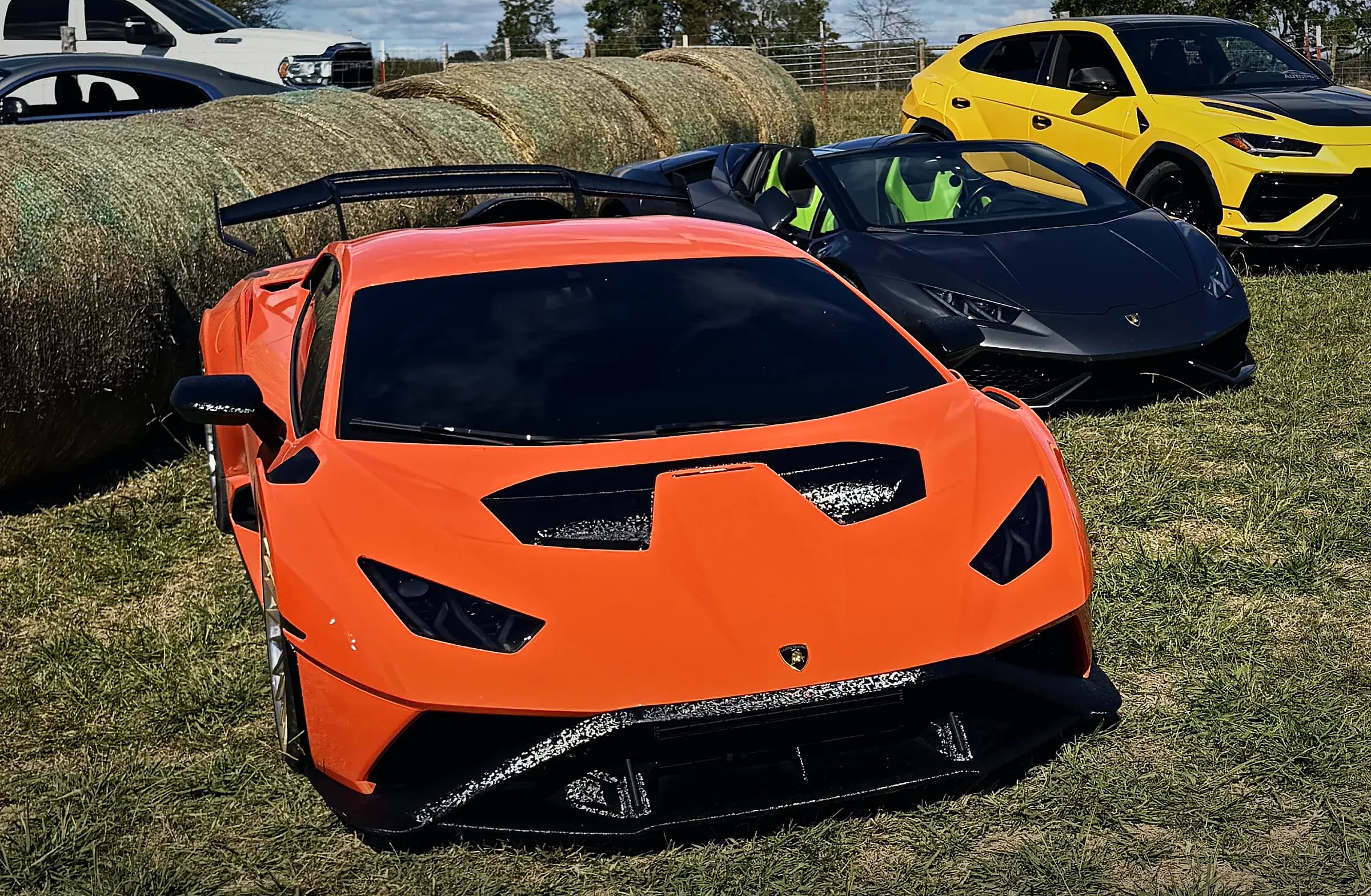 The orange Huracán STO up close on the grass at the ranch, its carbon wing and hood vents, a black Lamborghini and a yellow Urus behind it