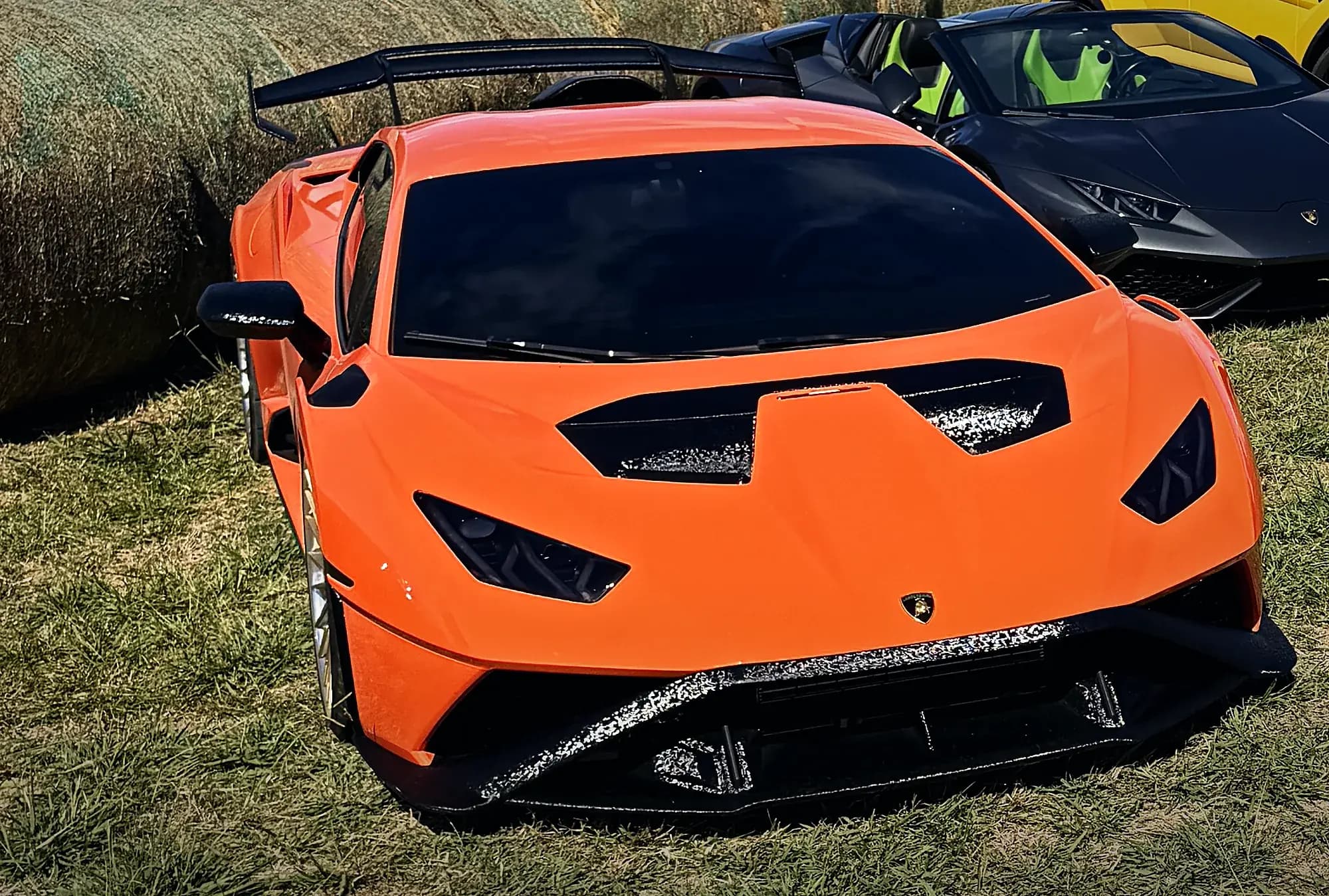 The orange Lamborghini Huracán STO filling the frame on the grass at Rancho Jaramillo, its carbon wing and hood vents, round hay bales and a black Lamborghini behind it