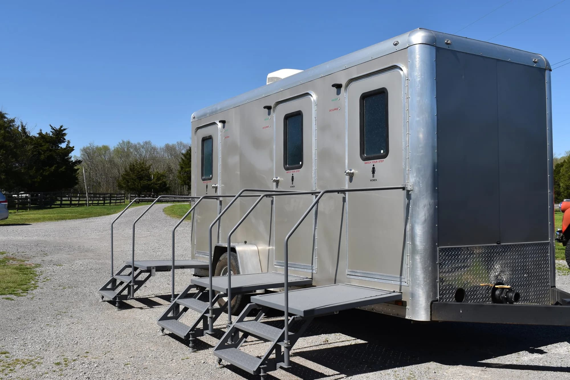 A silver three-stall restroom trailer with steps and a handrail at each door, parked on a gravel lane by a board fence