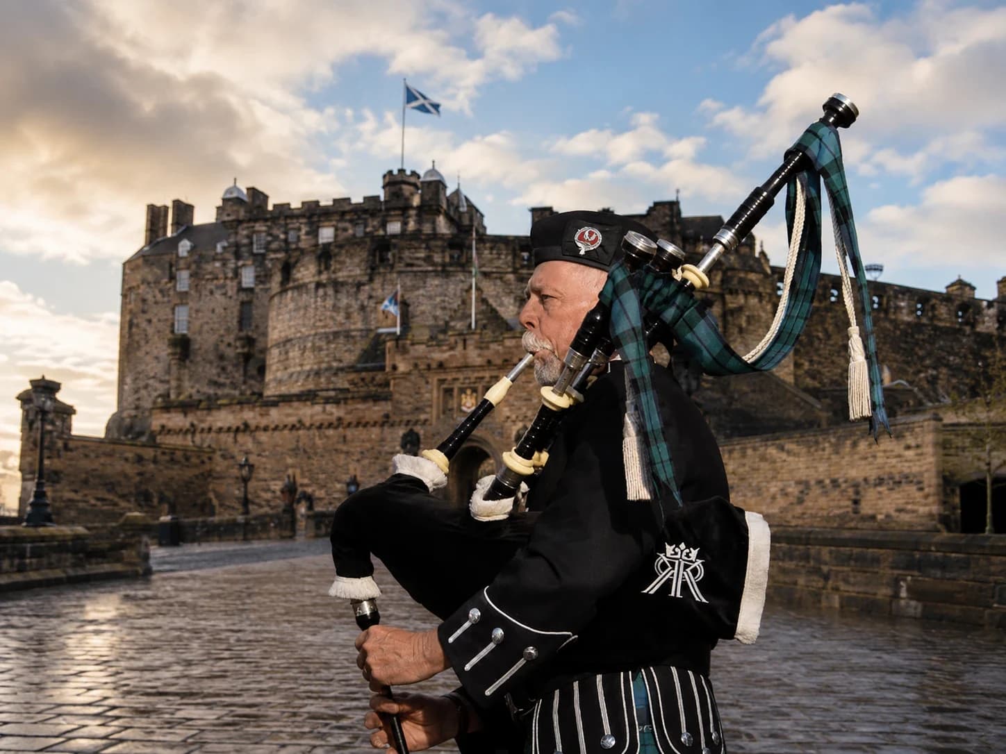 Richard Katz in Highland dress plays the bagpipes in front of Edinburgh Castle, a Saltire flying above it