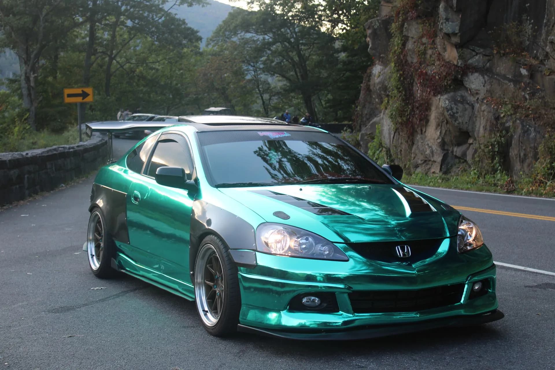 A green chrome coupe with wide fenders, a carbon hood and a carbon wing on a bend of a mountain road